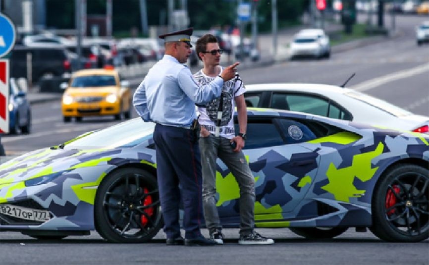 Evil Corp leader Maksim Yakubets talks to a Russian policeman in front of his Lamborghini with the license plate “VOR” (lit. “THIEF”)