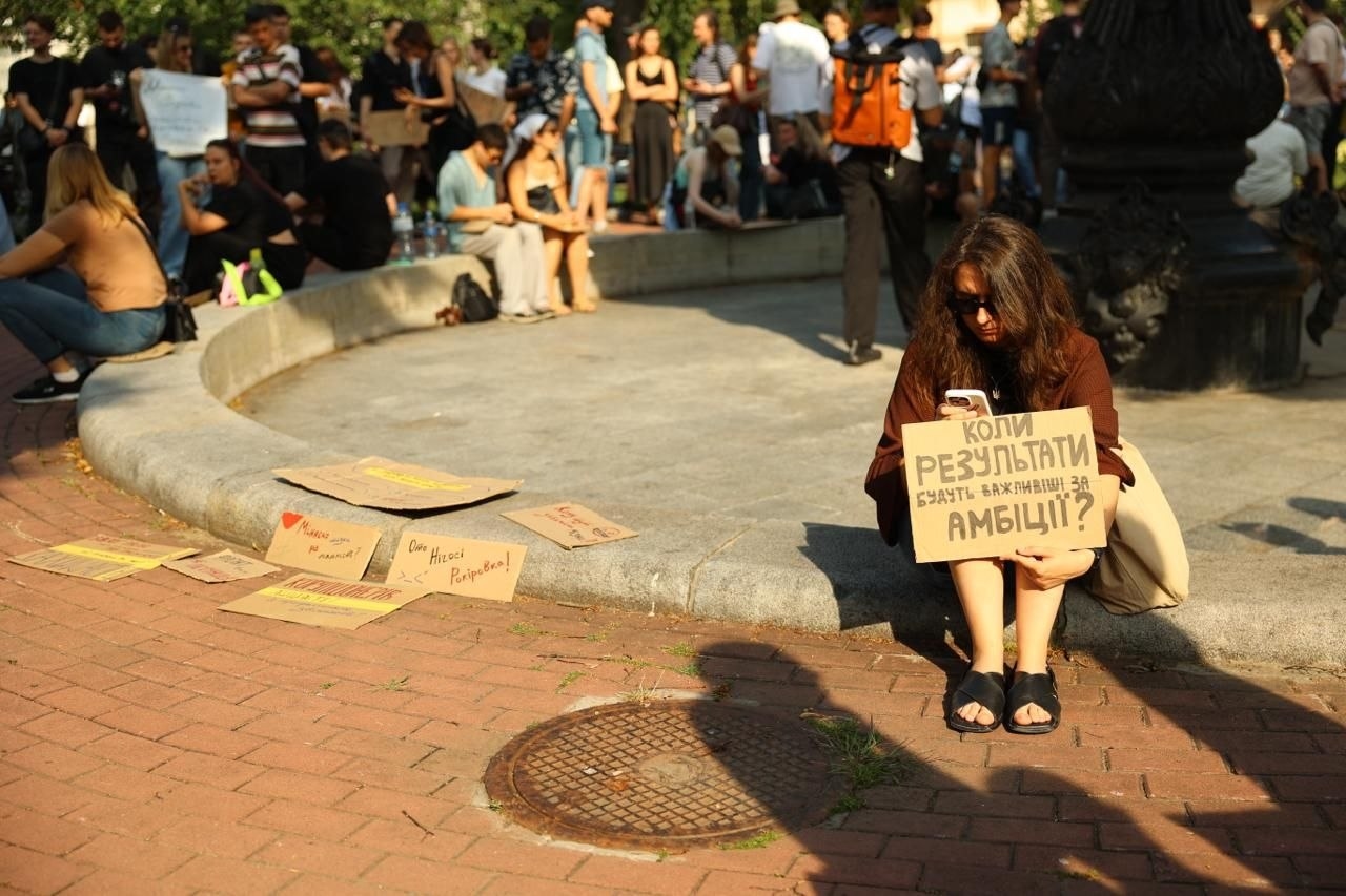 A woman holding a placard reading “When will results be more important than ambitions?”