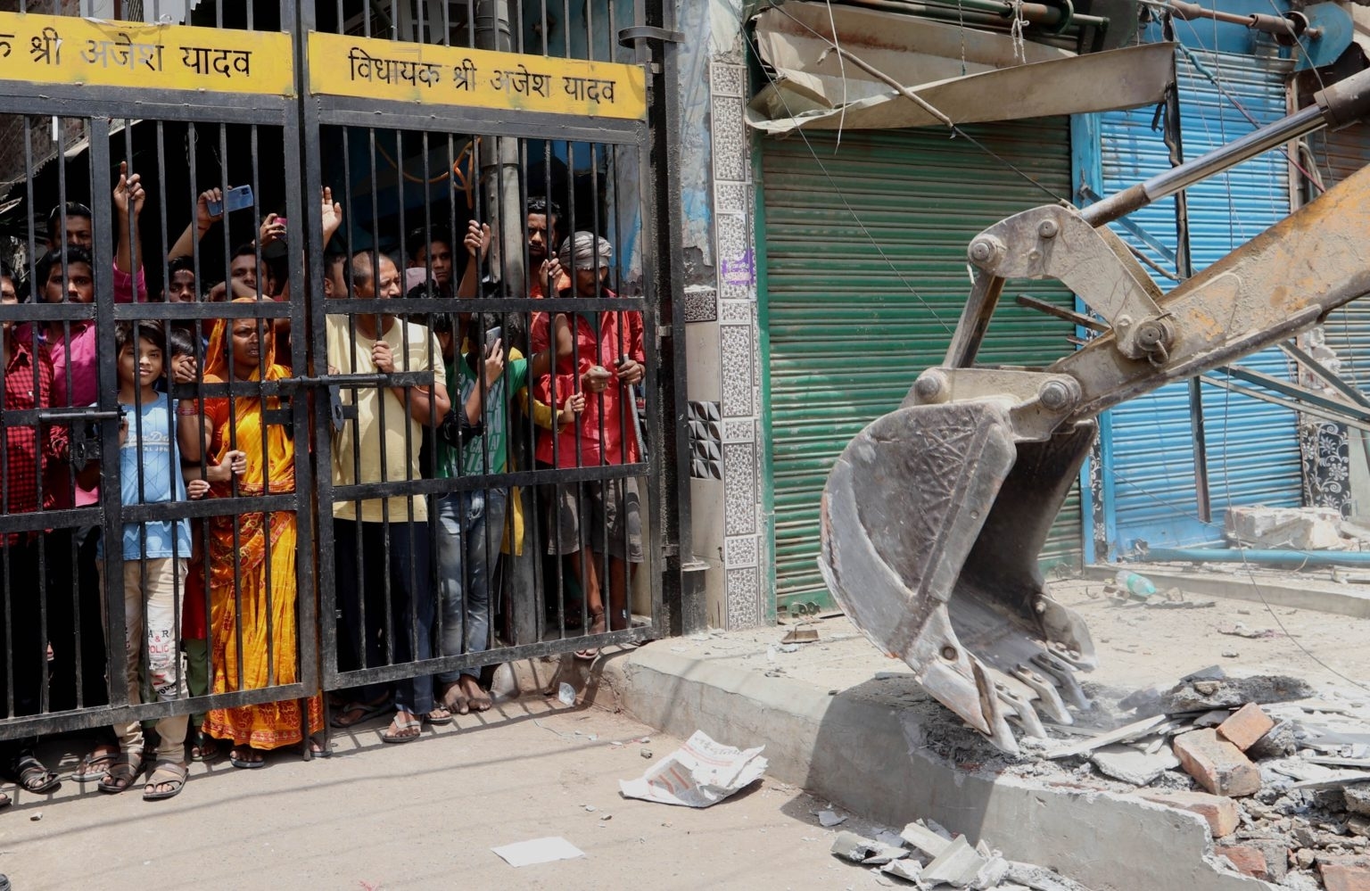 Local residents stand behind an iron gate watching a bulldozer demolish a structure in Jahangirpuri, Delhi, India